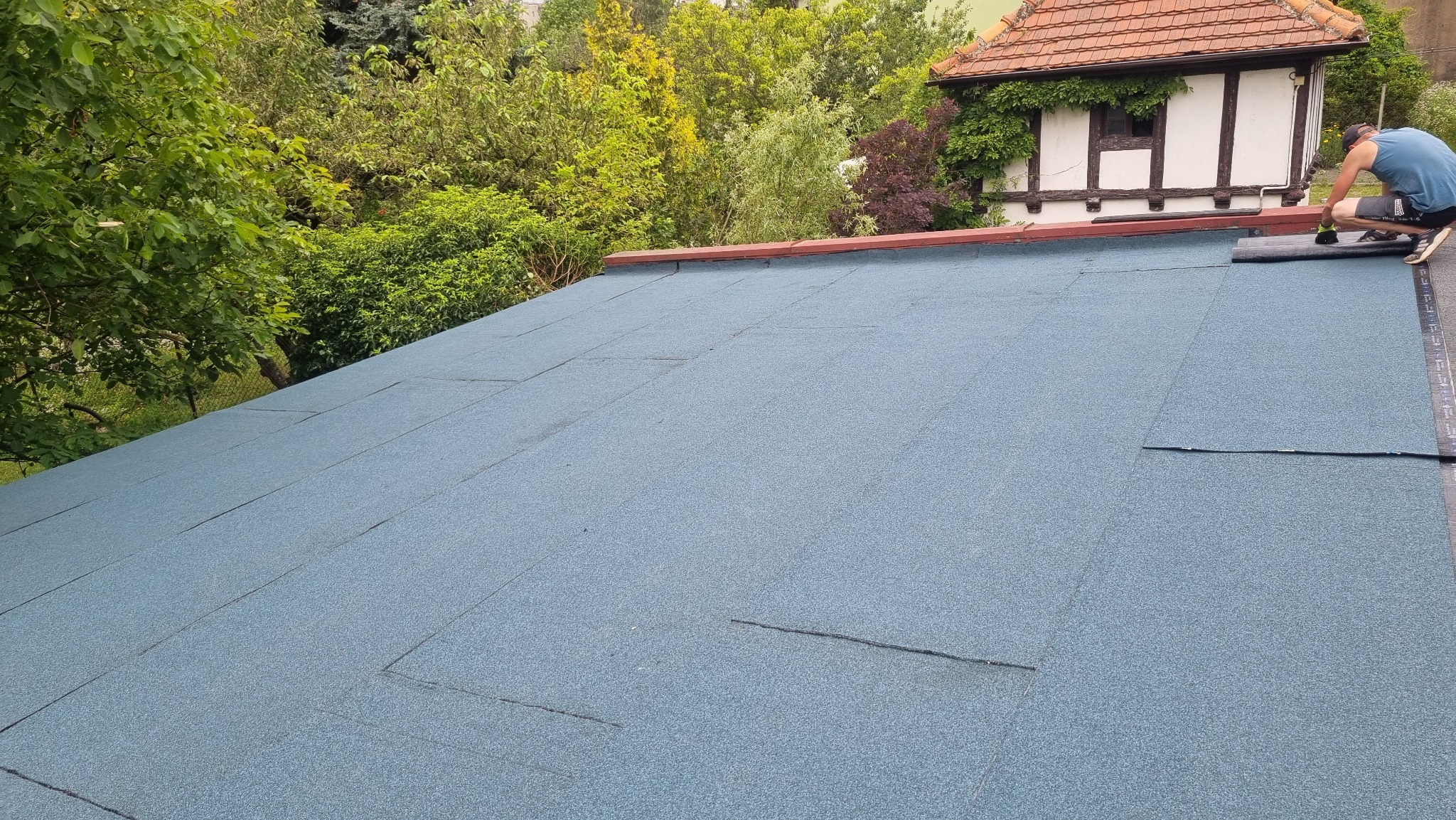 Pracownik w trakcie układania papy termozgrzewalnej na płaskim dachu; widoczny fragment dachu z nową, niebieską izolacją, narzędzia pracy i otoczenie z zielenią.