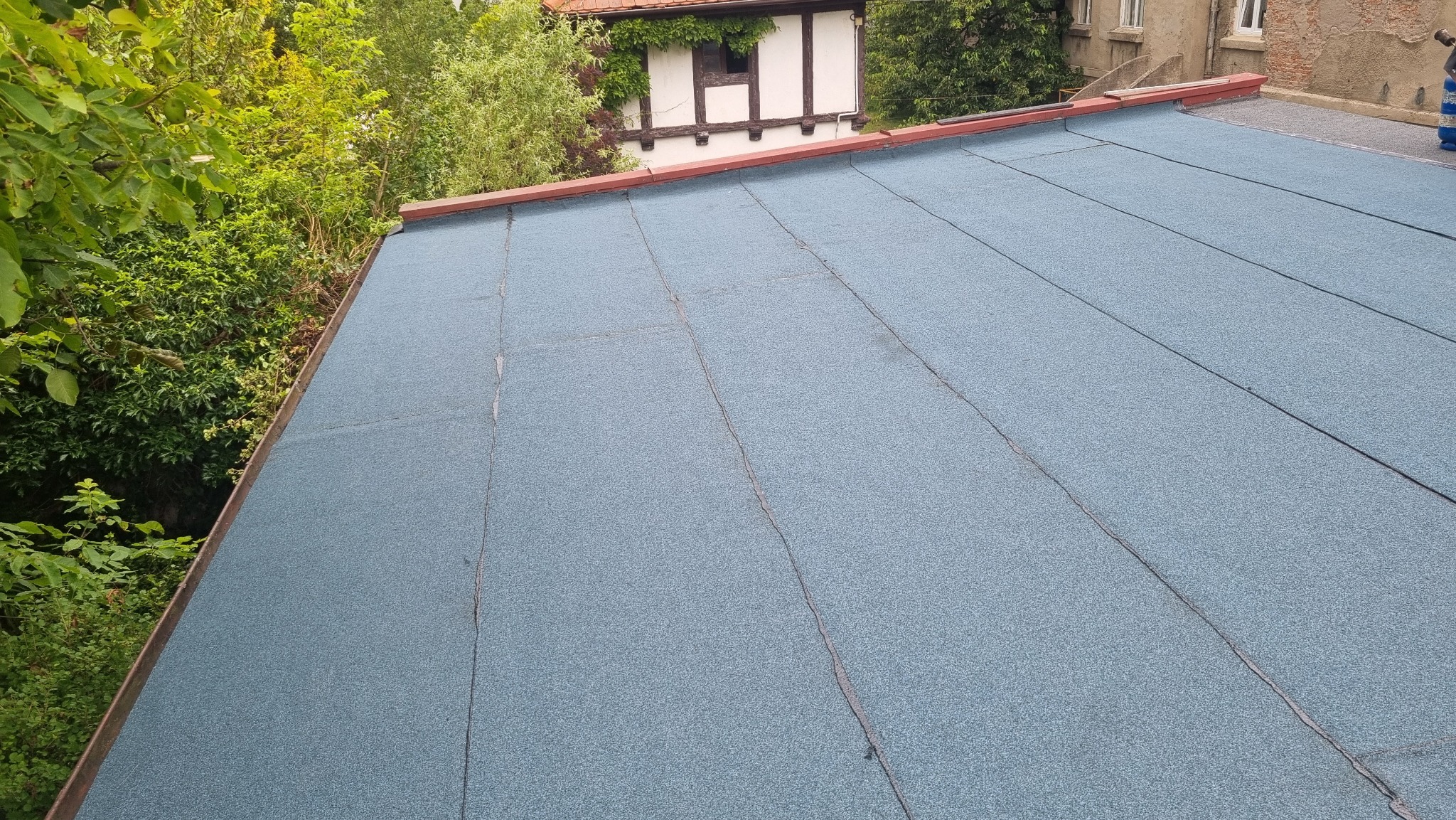 Płaski dach pokryty papą termozgrzewalną w kolorze szaroniebieskim, widok z góry, na tle budynek z muru pruskiego i zieleń.