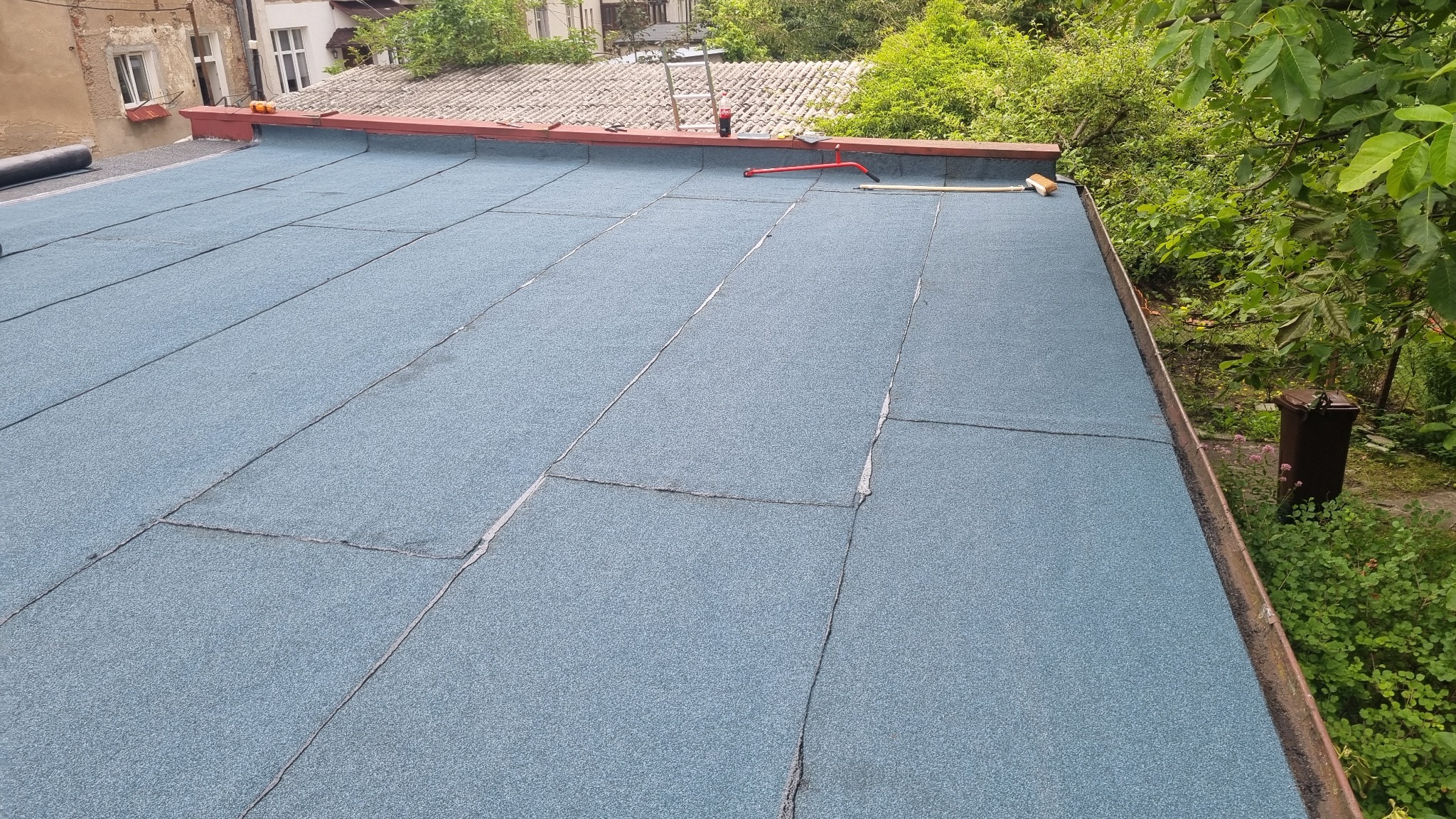 Dach pokryty niebieską papą termozgrzewalną, widoczne narzędzia dekarskie i fragment otoczenia z zielenią.