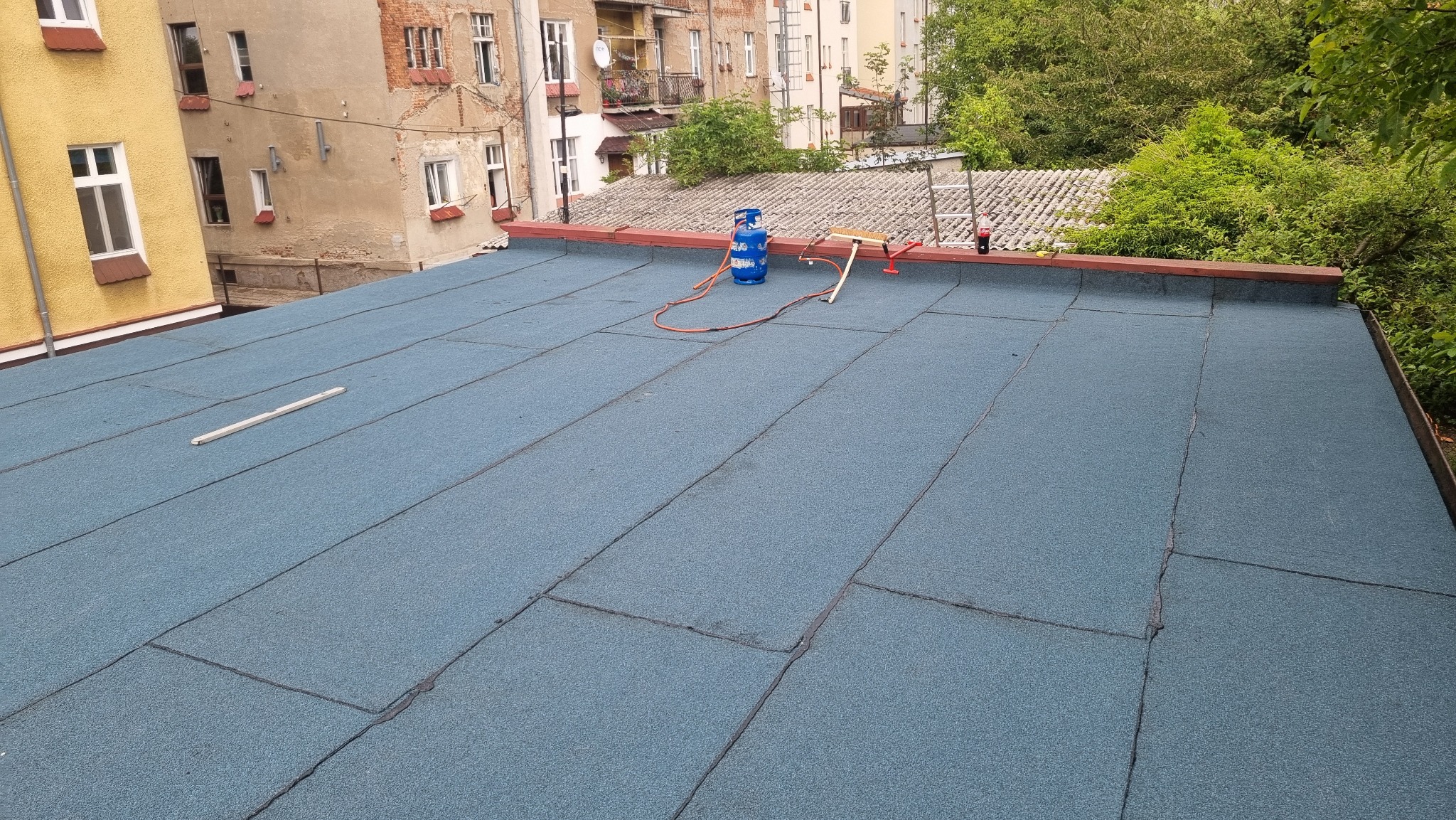 Niebieska papa termozgrzewalna na płaskim dachu, widoczny butla gazowa, palnik dekarski, drabina oraz zabudowa miejska w tle.