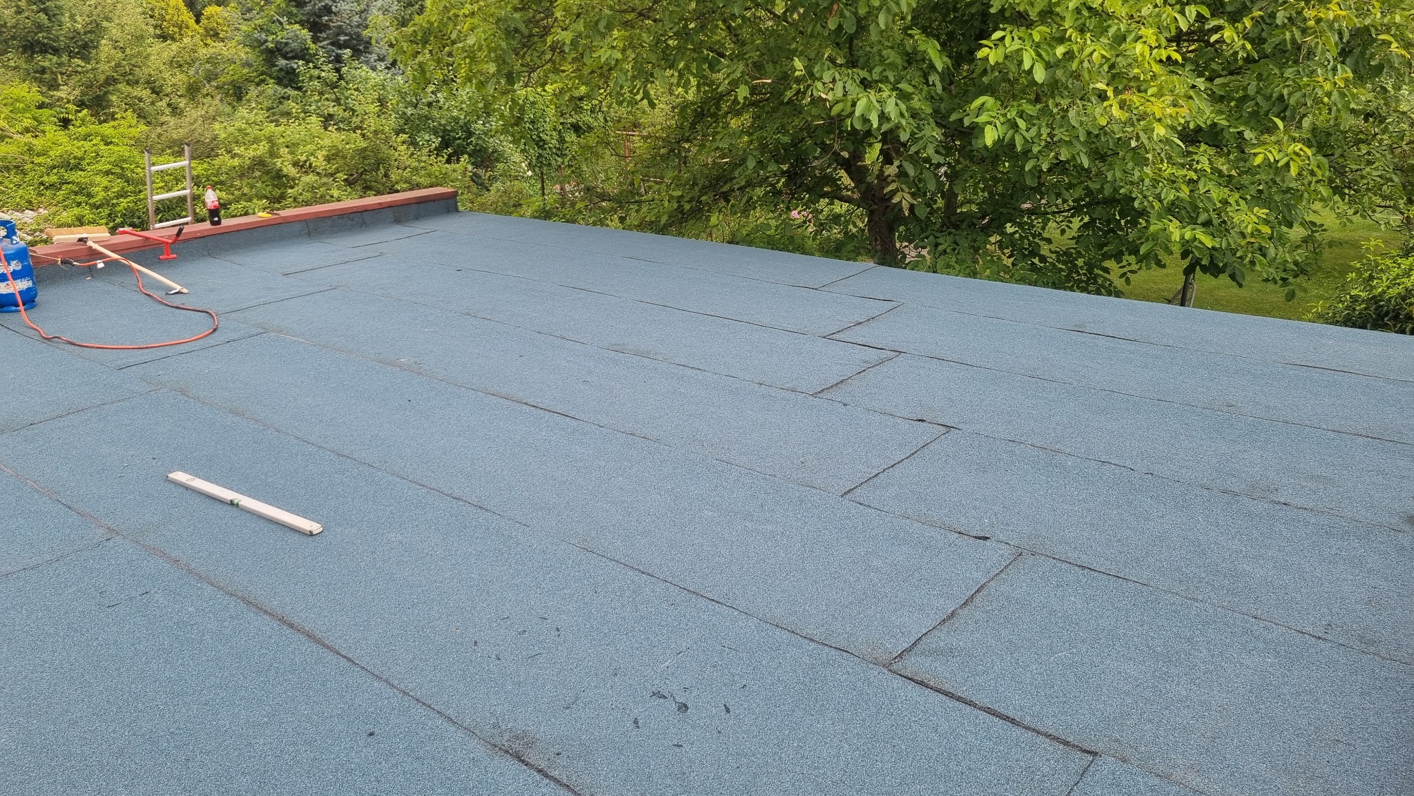 Płaski dach pokryty papą termozgrzewalną w odcieniach szarości i błękitu, widoczne narzędzia dekarskie, butla gazowa, drabina i zieleń w tle.