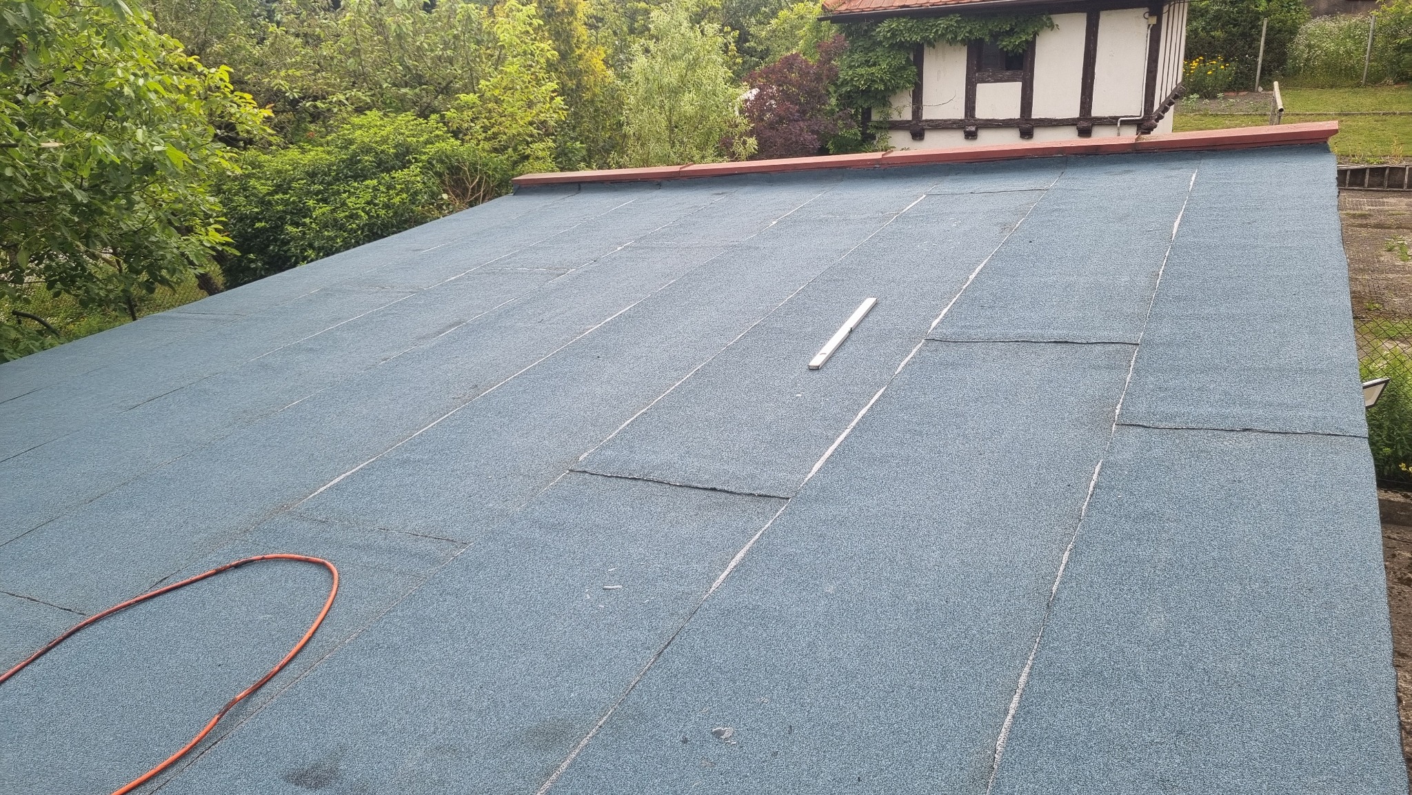 Widok z góry na płaski dach pokryty papą termozgrzewalną w kolorze niebiesko-szarym, z widocznymi łączeniami i fragmentem pomarańczowego kabla oraz poziomicą w centralnej części, w tle budynek...