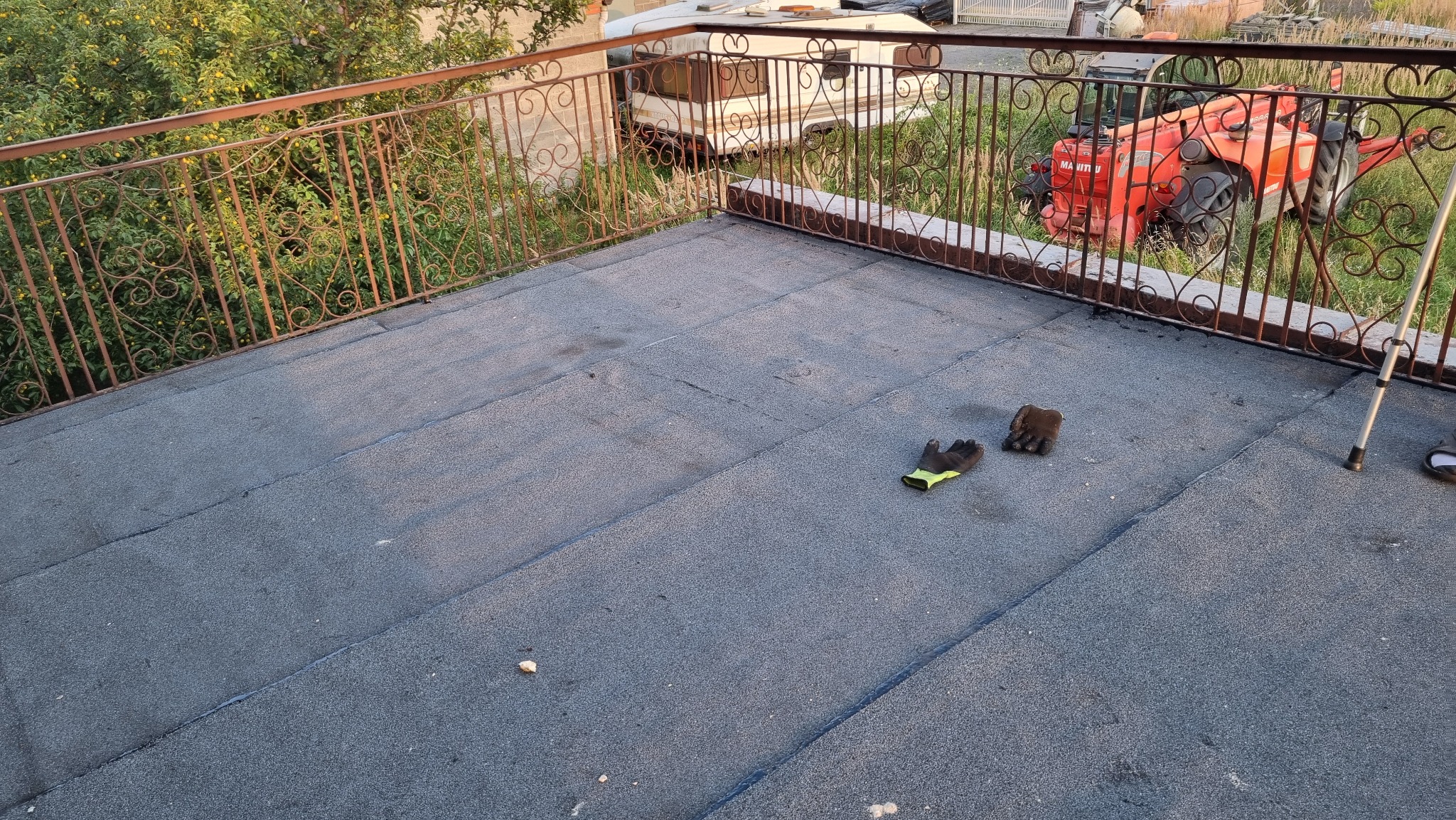 Płaski dach pokryty papą, z widocznymi łączeniami, na którym leżą robocze rękawice; w tle metalowa balustrada, zieleń i maszyna budowlana.