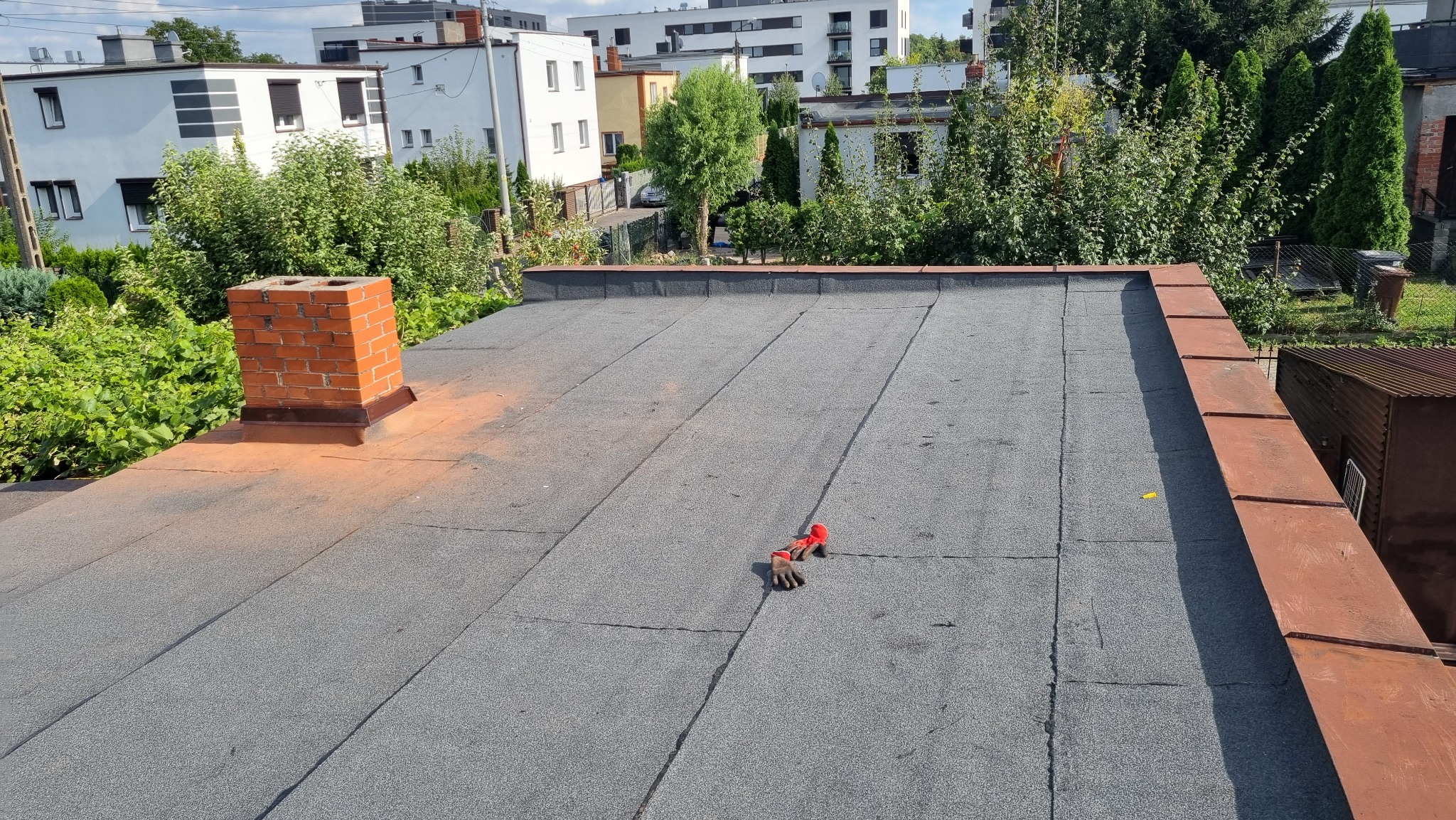 Płaski dach pokryty papą termozgrzewalną z ceglanym kominem i parą roboczych rękawic leżących na powierzchni; w tle budynki mieszkalne i zieleń.