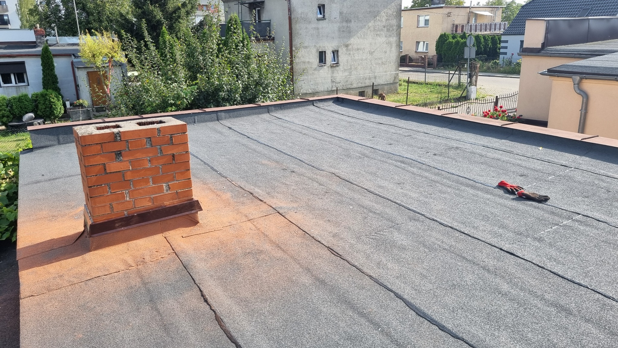 Płaski dach pokryty papą, z widocznym kominem z cegły i parą rękawic roboczych na powierzchni dachu.