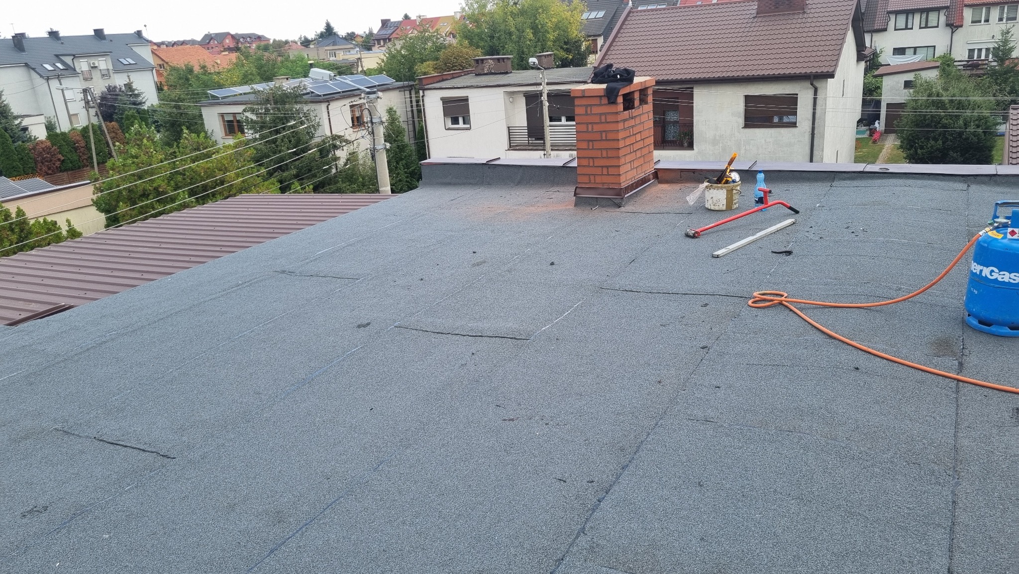 Szary, płaski dach pokryty papą termozgrzewalną z widocznym kominem z czerwonej cegły, butlą gazową, narzędziami i akcesoriami dekarskimi w słoneczny dzień.