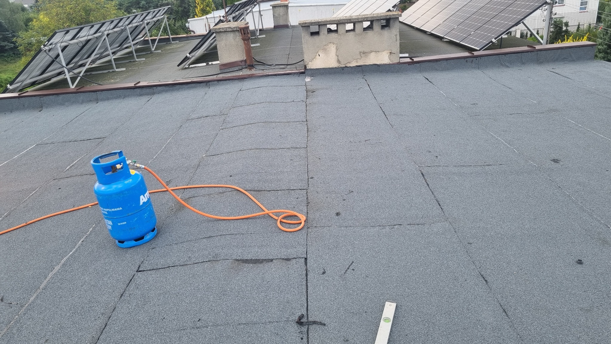 Niedokończone krycie dachu papą termozgrzewalną, widoczna butla gazowa z palnikiem i poziomica, w tle panele solarne i kominy.