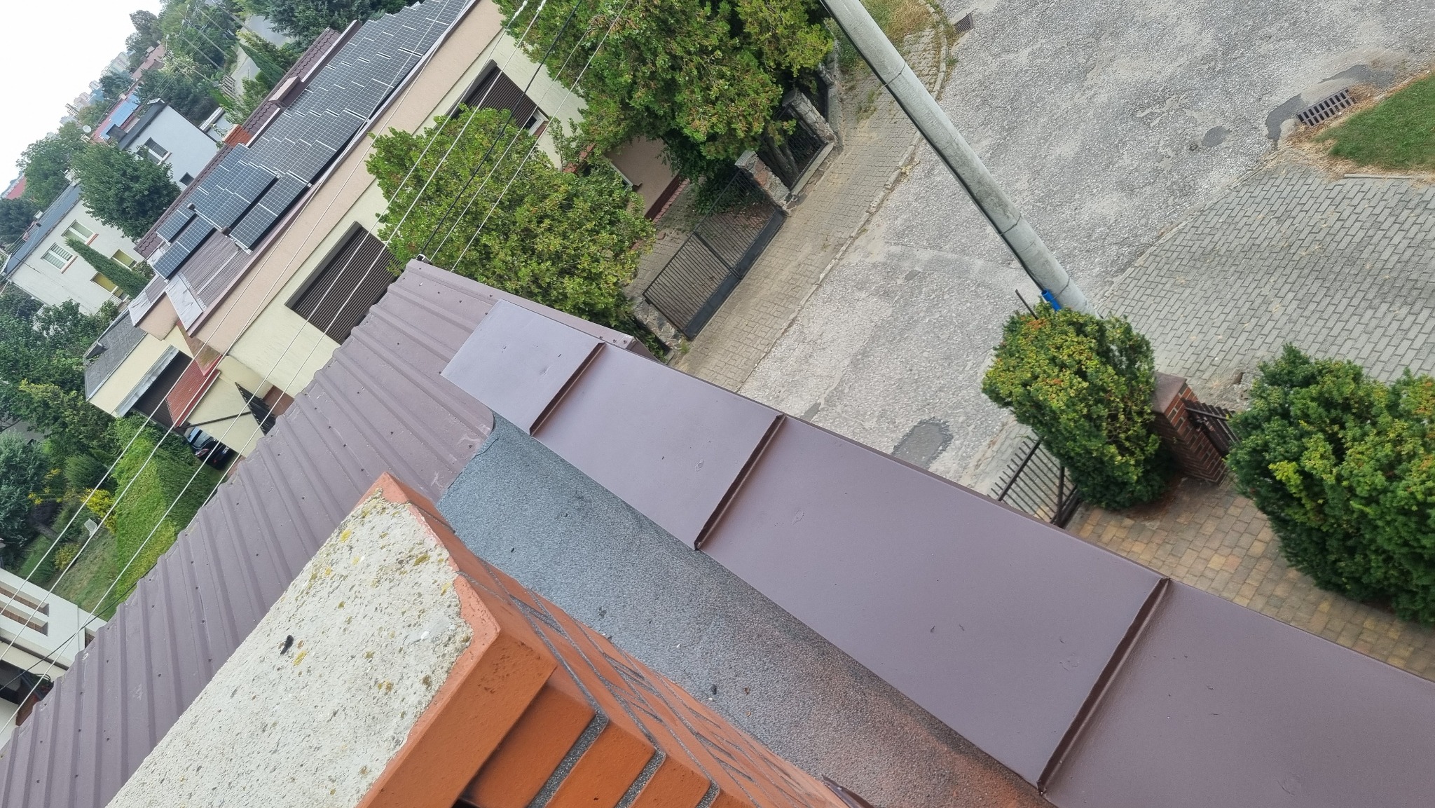 Widok z góry na brązowy dach z blachy trapezowej, fragment komina z cegły i betonowych elementów, w tle budynki mieszkalne z panelami słonecznymi i zielenią.