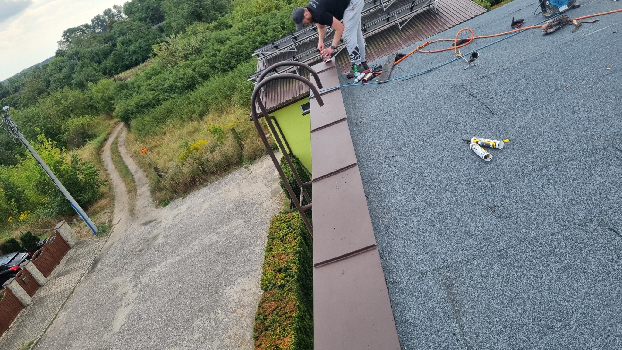 Pracownik w czarnej koszulce i szarych spodniach na dachu pokrytym papą, zabezpiecza obróbkę blacharską przy użyciu silikonu dekarskiego; w tle drabina dekarska i zielone drzewa.