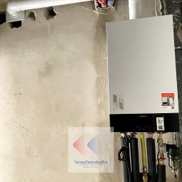 Termotechnika.Pro Rostysław Lisowski - Nowoczesny, biały piec gazowy Viessmann zamontowany na surowej ścianie. Widoczne rury instalacyjne, w tym biała rura odprowadzająca spaliny. Instalacja w trakcie prac wykończeniowych.
