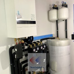 Termotechnika.Pro Rostysław Lisowski - Nowoczesna instalacja grzewcza z piecem gazowym Panasonic, zbiornikiem owiniętym folią i czarnymi rurami. Widoczne manometry i zawory. Elementy instalacji pod sufitem.