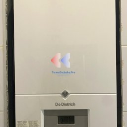 Termotechnika.Pro Rostysław Lisowski - Biały, wiszący piec gazowy De Dietrich z wyświetlaczem i naklejką serwisową, zamontowany na tle jasnych płytek. Widoczne przyłącza gazowe i hydrauliczne.