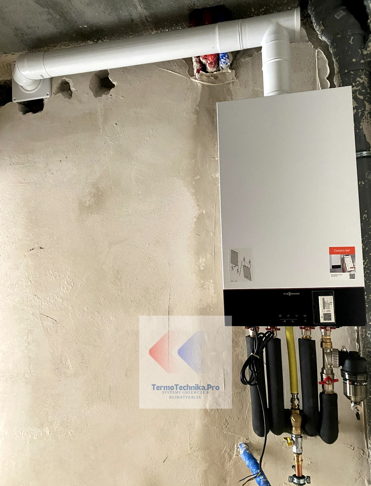 Nowoczesny, biały piec gazowy Viessmann zamontowany na surowej ścianie. Widoczne rury instalacyjne, w tym biała rura odprowadzająca spaliny. Instalacja w trakcie prac wykończeniowych.