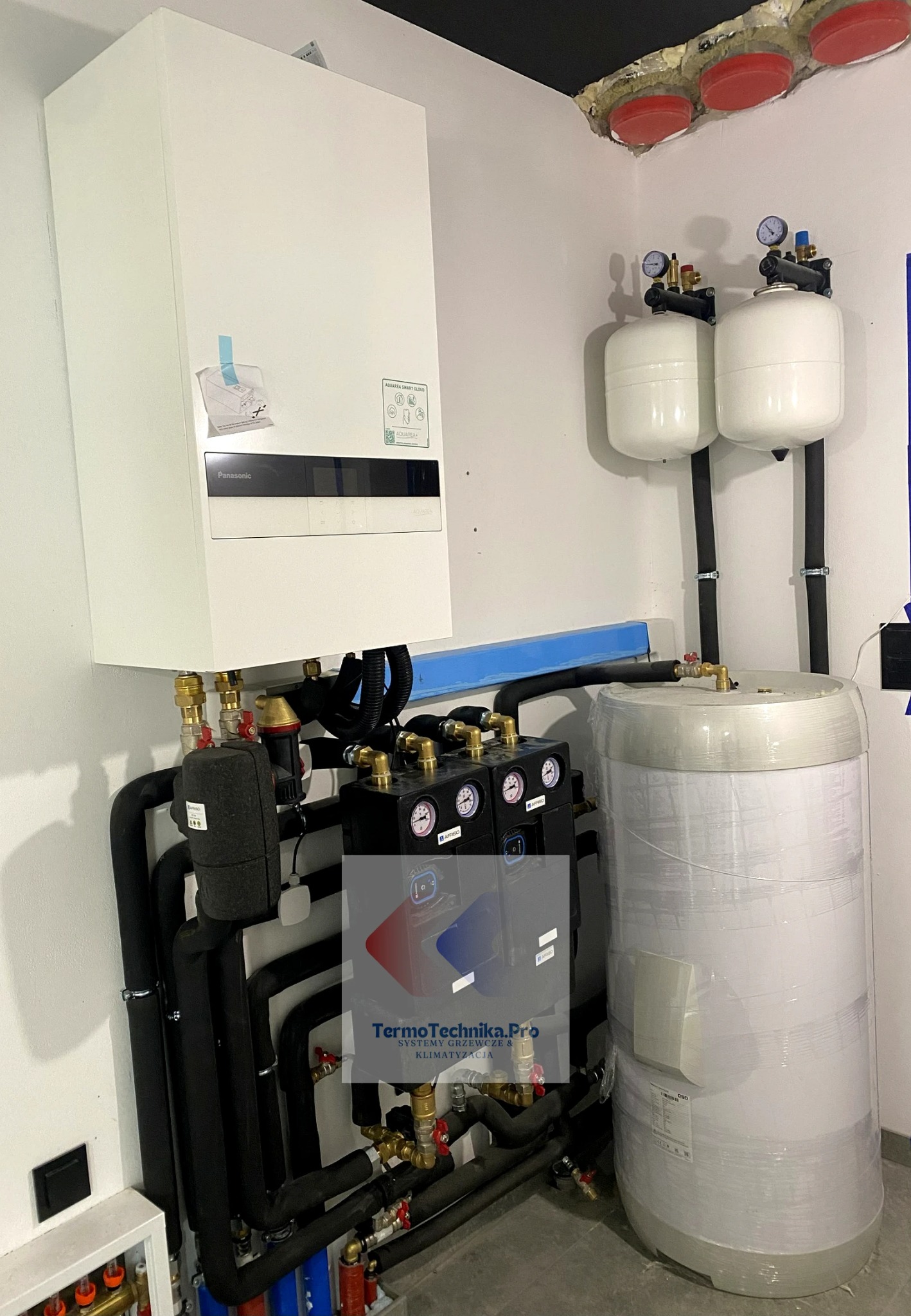 Nowoczesna instalacja grzewcza z piecem gazowym Panasonic, zbiornikiem owiniętym folią i czarnymi rurami. Widoczne manometry i zawory. Elementy instalacji pod sufitem.