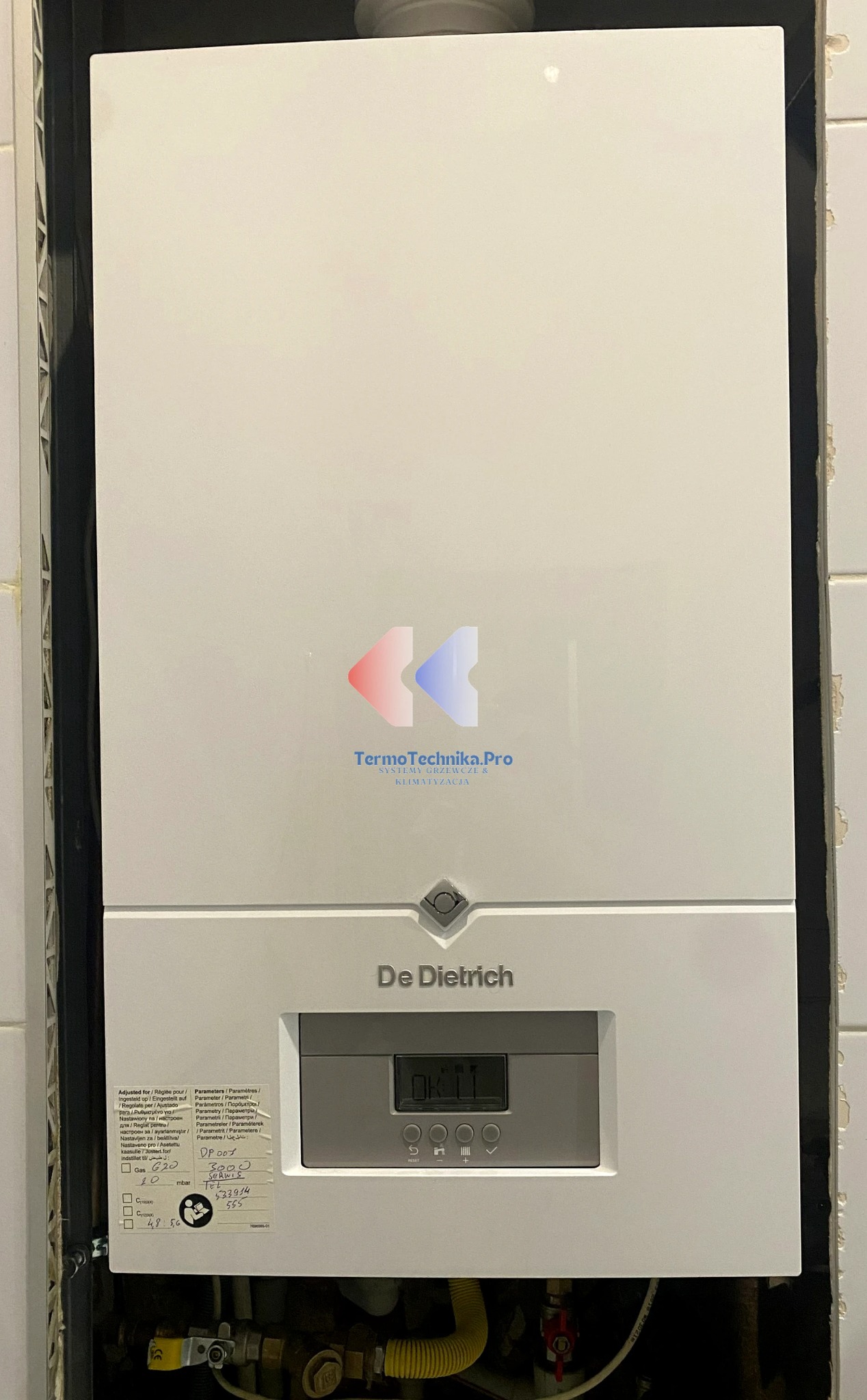Biały, wiszący piec gazowy De Dietrich z wyświetlaczem i naklejką serwisową, zamontowany na tle jasnych płytek. Widoczne przyłącza gazowe i hydrauliczne.
