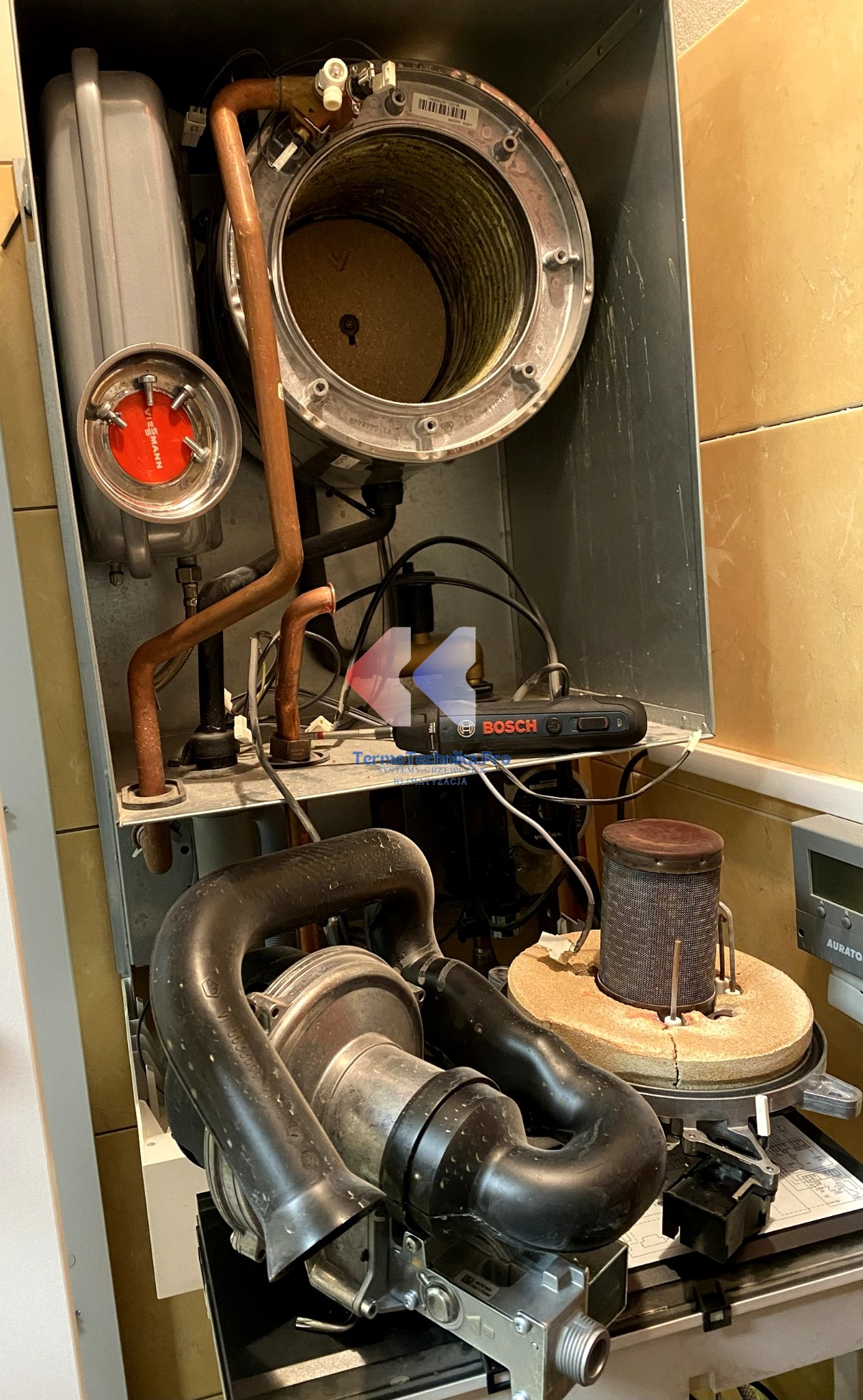 Wnętrze pieca gazowego Viessmann z widocznymi elementami instalacji, w tym miedzianymi rurami i czarnym wentylatorem. Widoczny osprzęt Bosch.