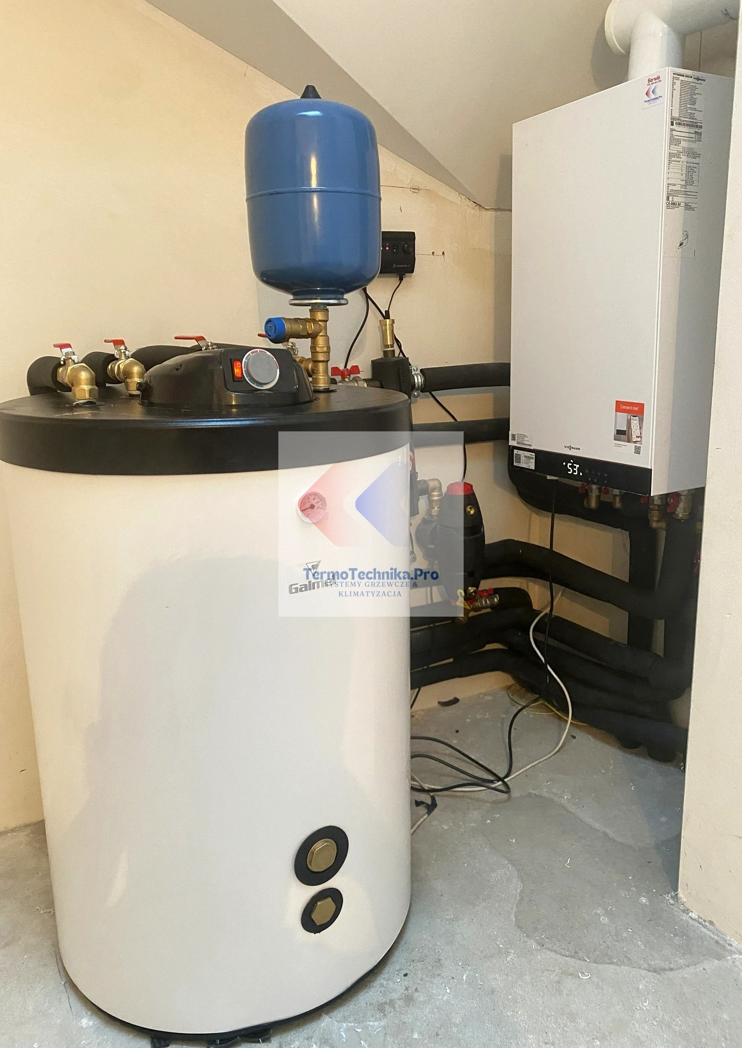 Biały piec gazowy Viessmann z czarnymi rurami i zbiornikiem wyrównawczym w kotłowni. Widoczne przyłącza i elementy instalacji. Oświetlenie górne.