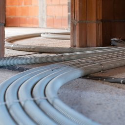 RekuRozwiązania - Szare, karbowane rury wentylacyjne prowadzone po surowej podłodze w budynku w trakcie budowy, widoczne ceglane ściany z pustaków ceramicznych.