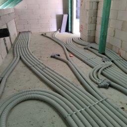 RekuRozwiązania - Szare, karbowane rury wentylacyjne ułożone na surowym betonowym podłożu w trakcie instalacji w budynku z białego pustaka, widoczne pomarańczowe zaślepki i zielony drewniany słup podtrzymujący strop.