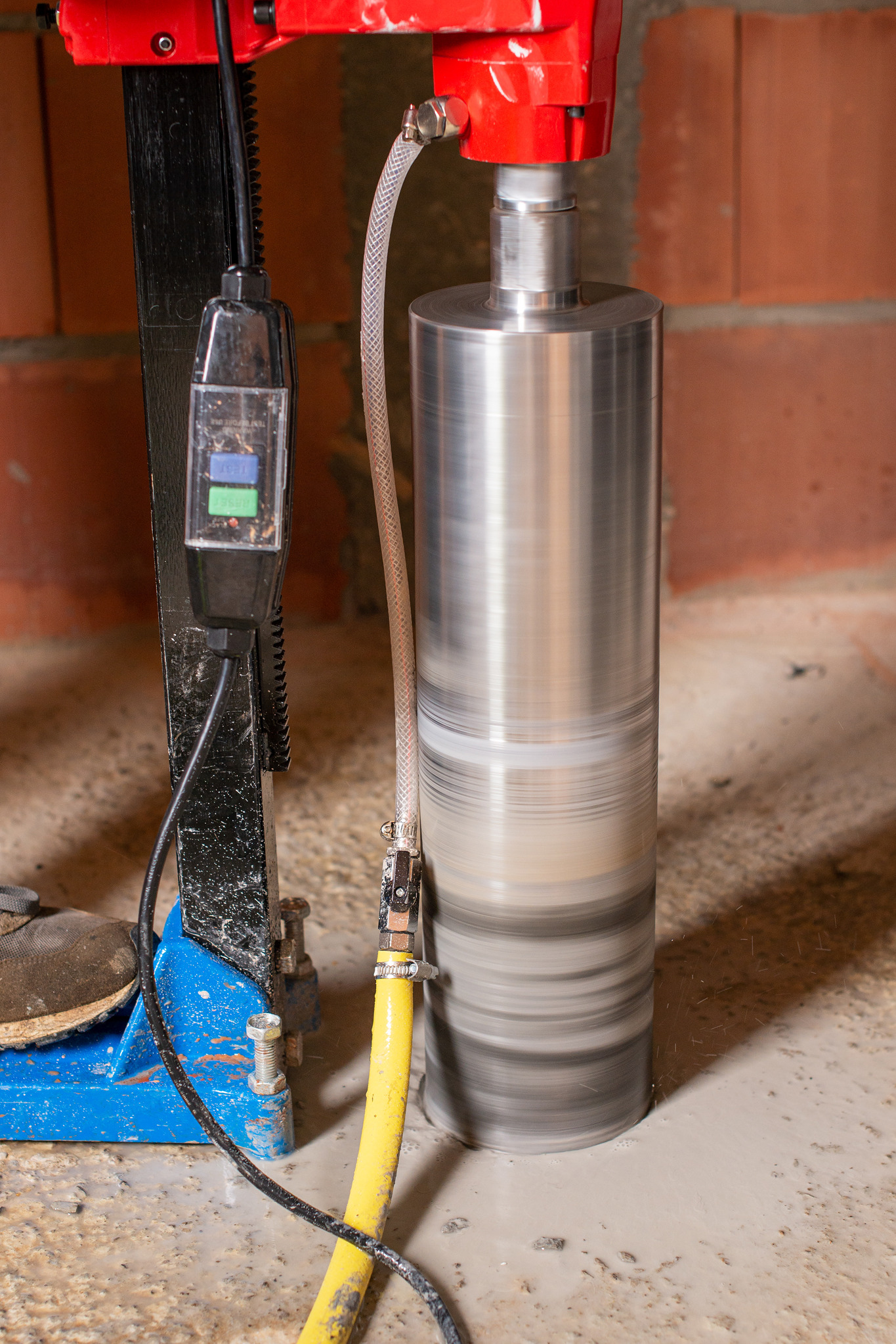 Wiertnica diamentowa w trakcie wiercenia otworu w betonowej podłodze, z widocznym systemem chłodzenia wodą i fragmentem ściany z cegły w tle.