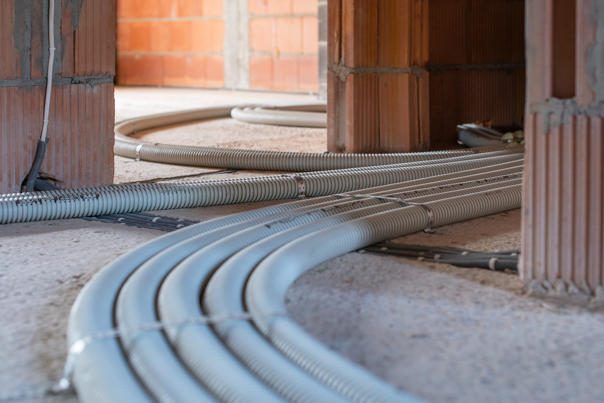 Szare, karbowane rury wentylacyjne prowadzone po surowej podłodze w budynku w trakcie budowy, widoczne ceglane ściany z pustaków ceramicznych.