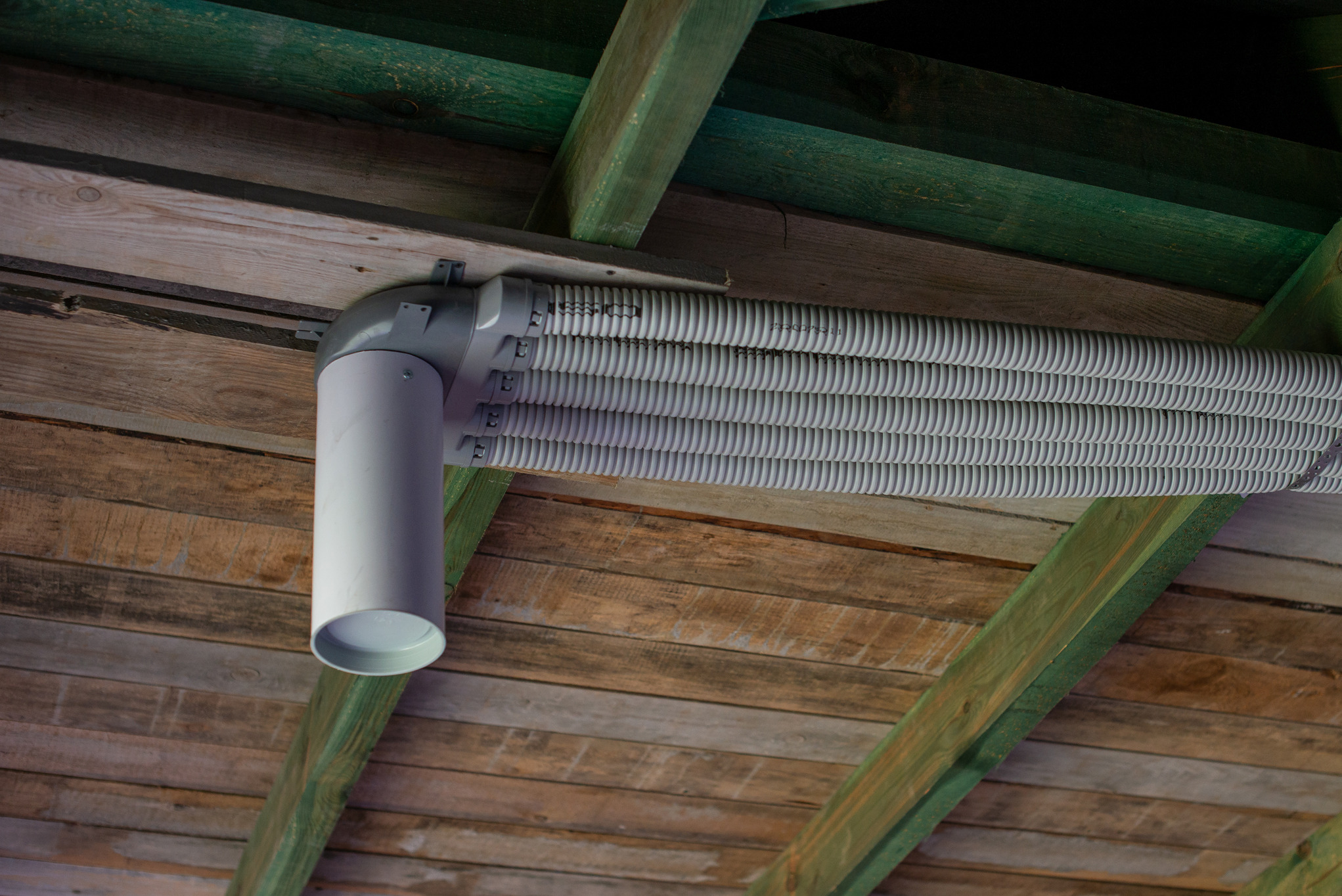 Cztery elastyczne, szare rury wentylacyjne zamontowane pod drewnianym stropem z zielonymi belkami nośnymi, widoczne z dołu.