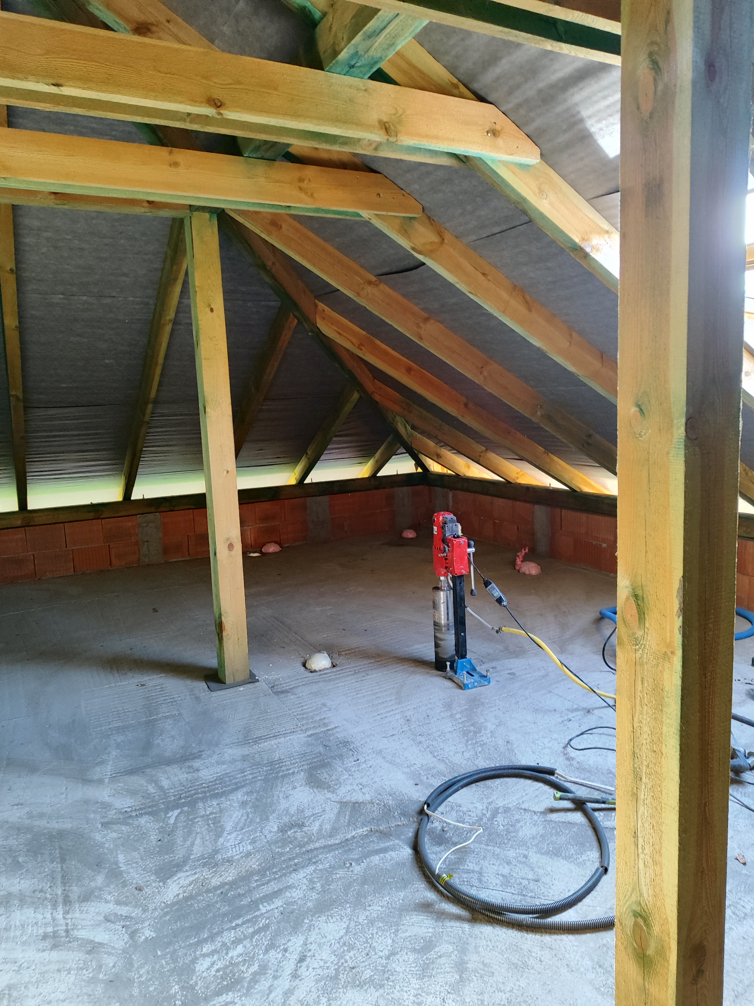 Wnętrze poddasza w trakcie prac instalacyjnych: widoczna konstrukcja drewniana dachu, ściany z czerwonej cegły, betonowa podłoga z widocznymi śladami po pracach, wiertnica diamentowa na podłodze...