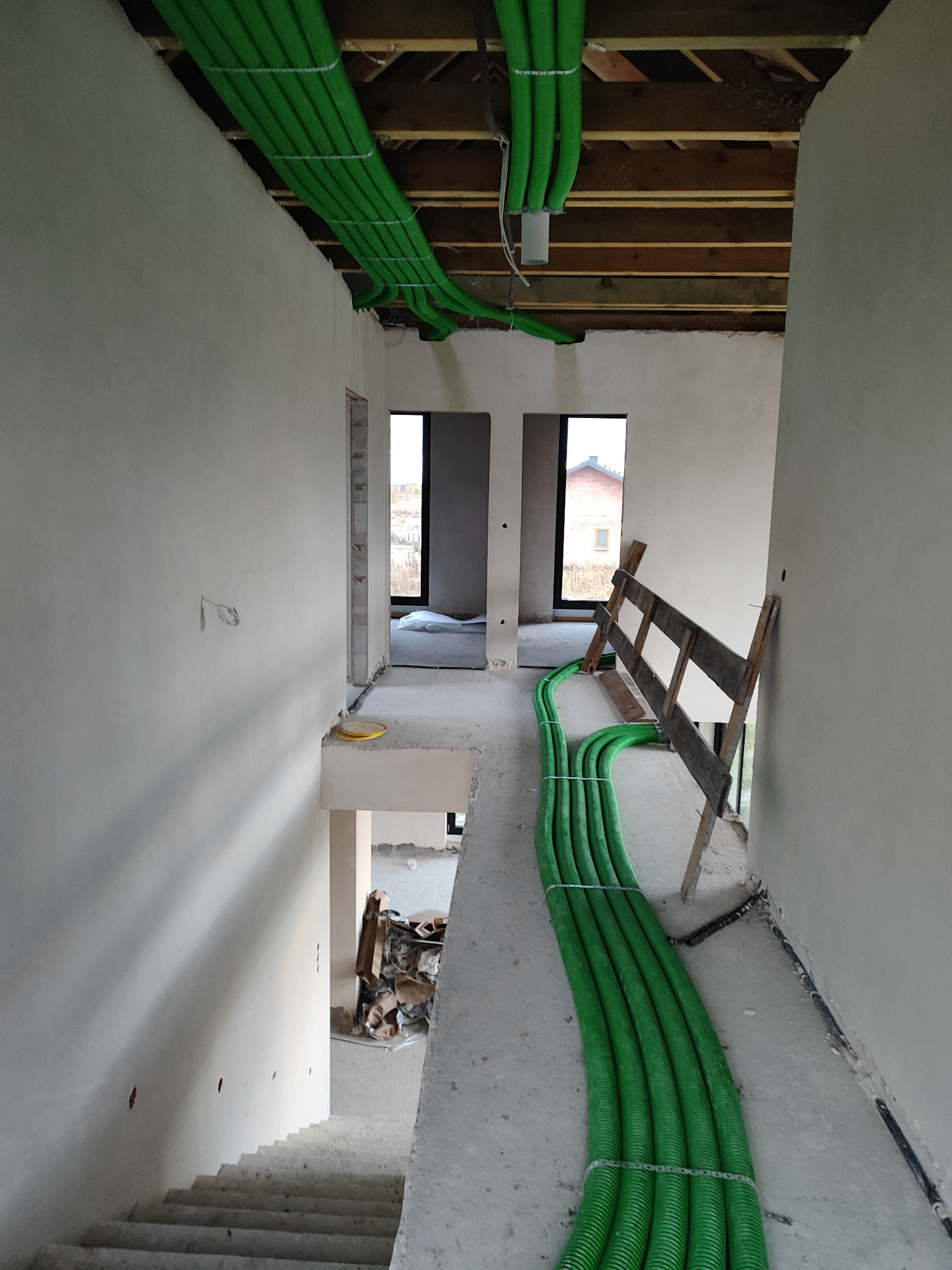 Wnętrze w trakcie montażu rekuperacji: wiązka zielonych rur wentylacyjnych przymocowana do sufitu i prowadzona wzdłuż krawędzi schodów, widoczne surowe ściany i drewniana konstrukcja stropu.