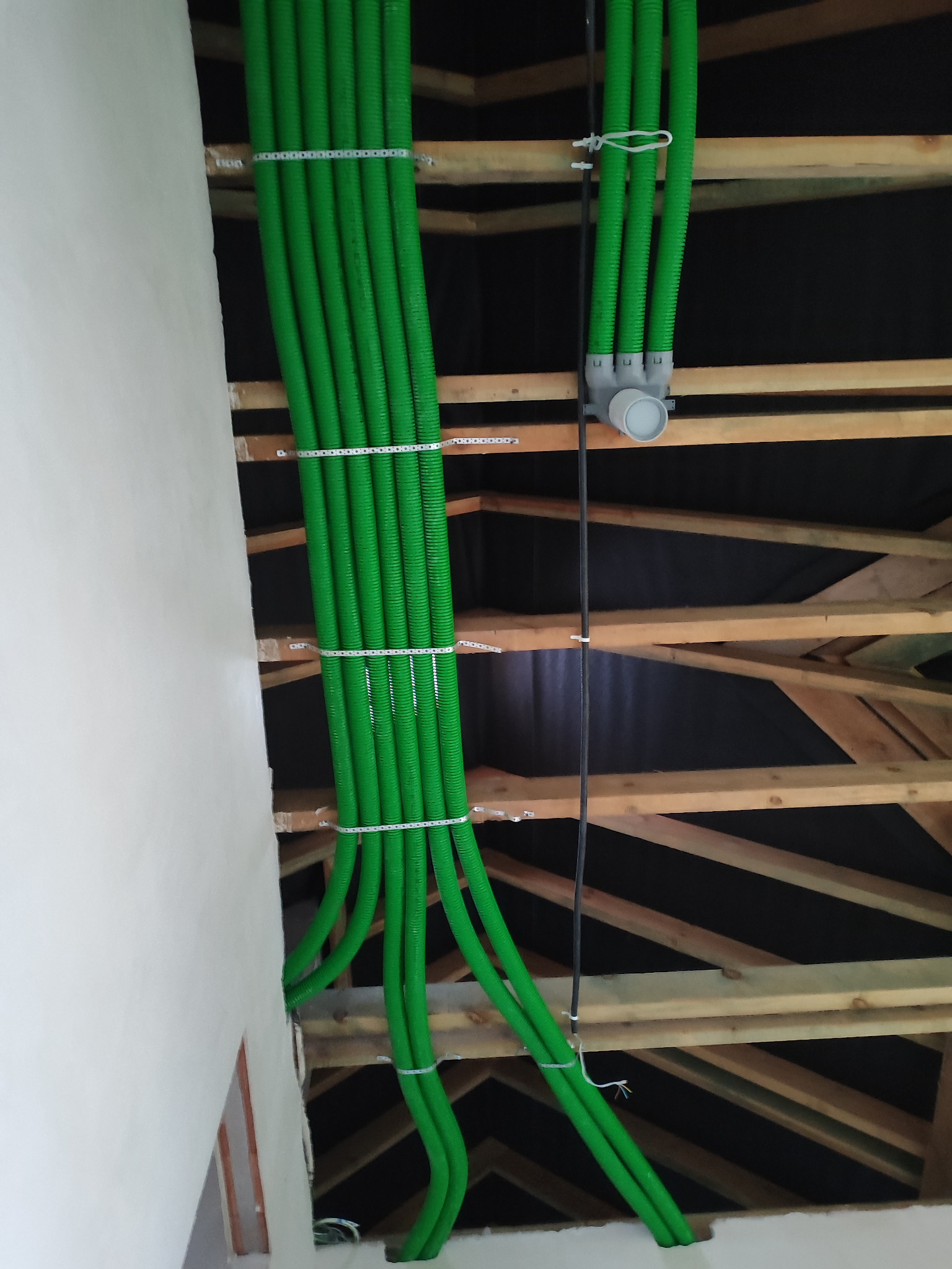 Zielone, karbowane rury wentylacyjne przymocowane do drewnianej konstrukcji dachu i ściany, widoczne elementy instalacji z szarym łącznikiem.