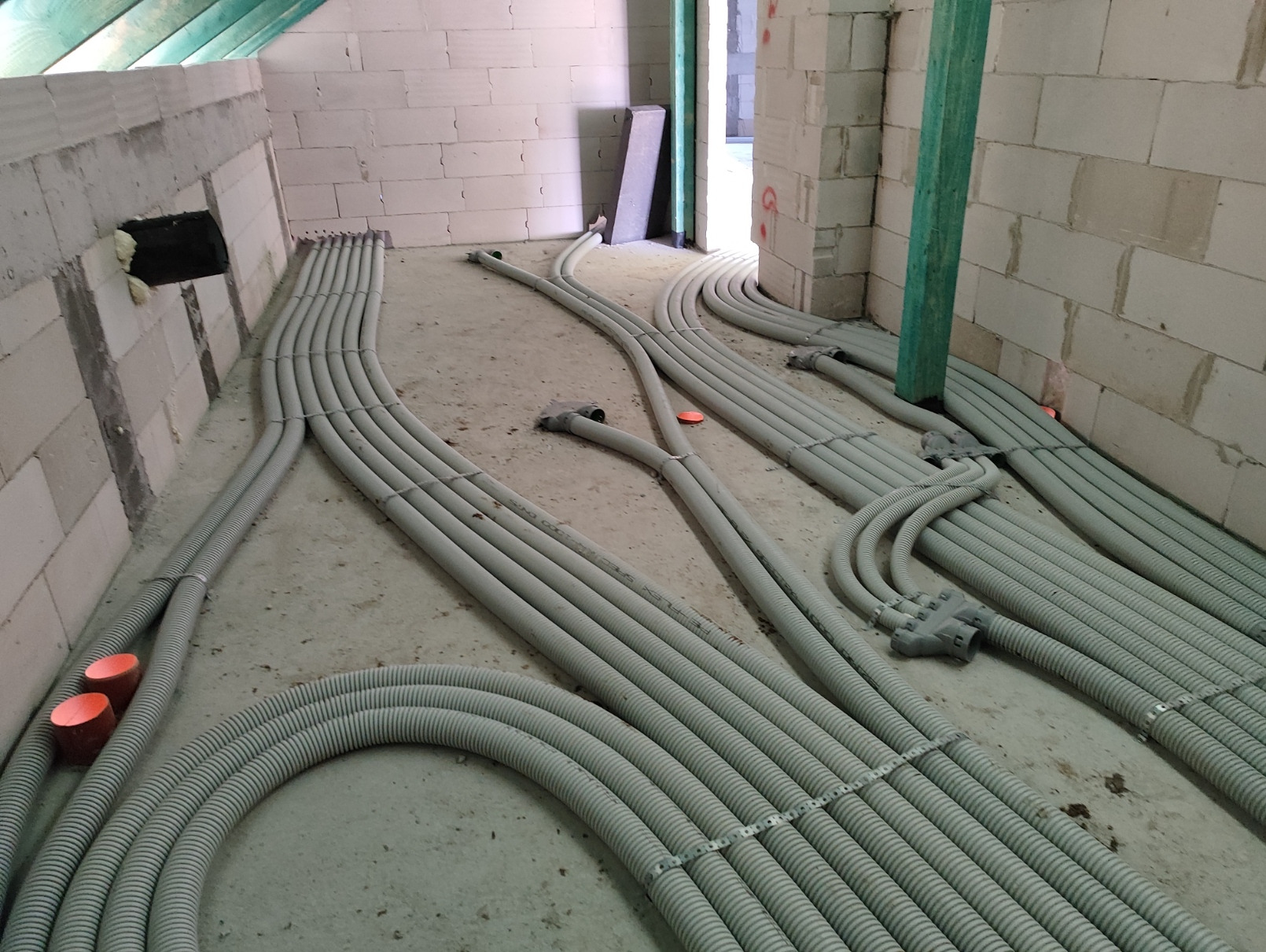 Szare, karbowane rury wentylacyjne ułożone na surowym betonowym podłożu w trakcie instalacji w budynku z białego pustaka, widoczne pomarańczowe zaślepki i zielony drewniany słup podtrzymujący strop.
