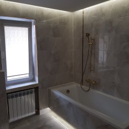 Frb Golański Dominik - Wykończona łazienka w szarościach z wanną, grzejnikiem pod oknem i oświetleniem LED. Płytki imitujące marmur z akcentami w kolorze złota.