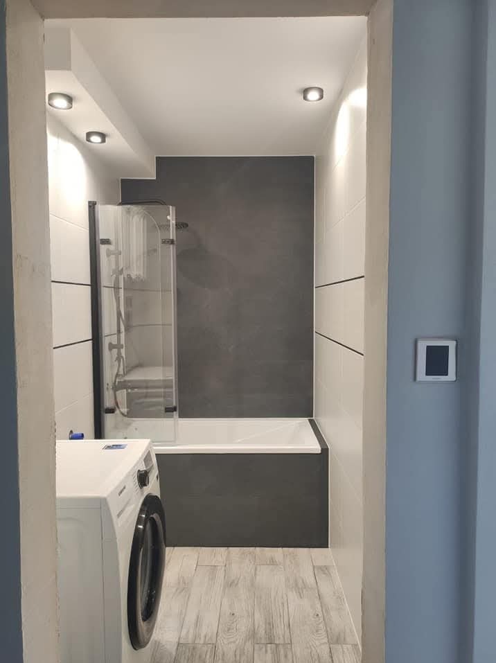 Wykończona łazienka z kabiną prysznicową nad wanną, biało-szare płytki, drewnopodobna podłoga i pralka. Widok z korytarza.