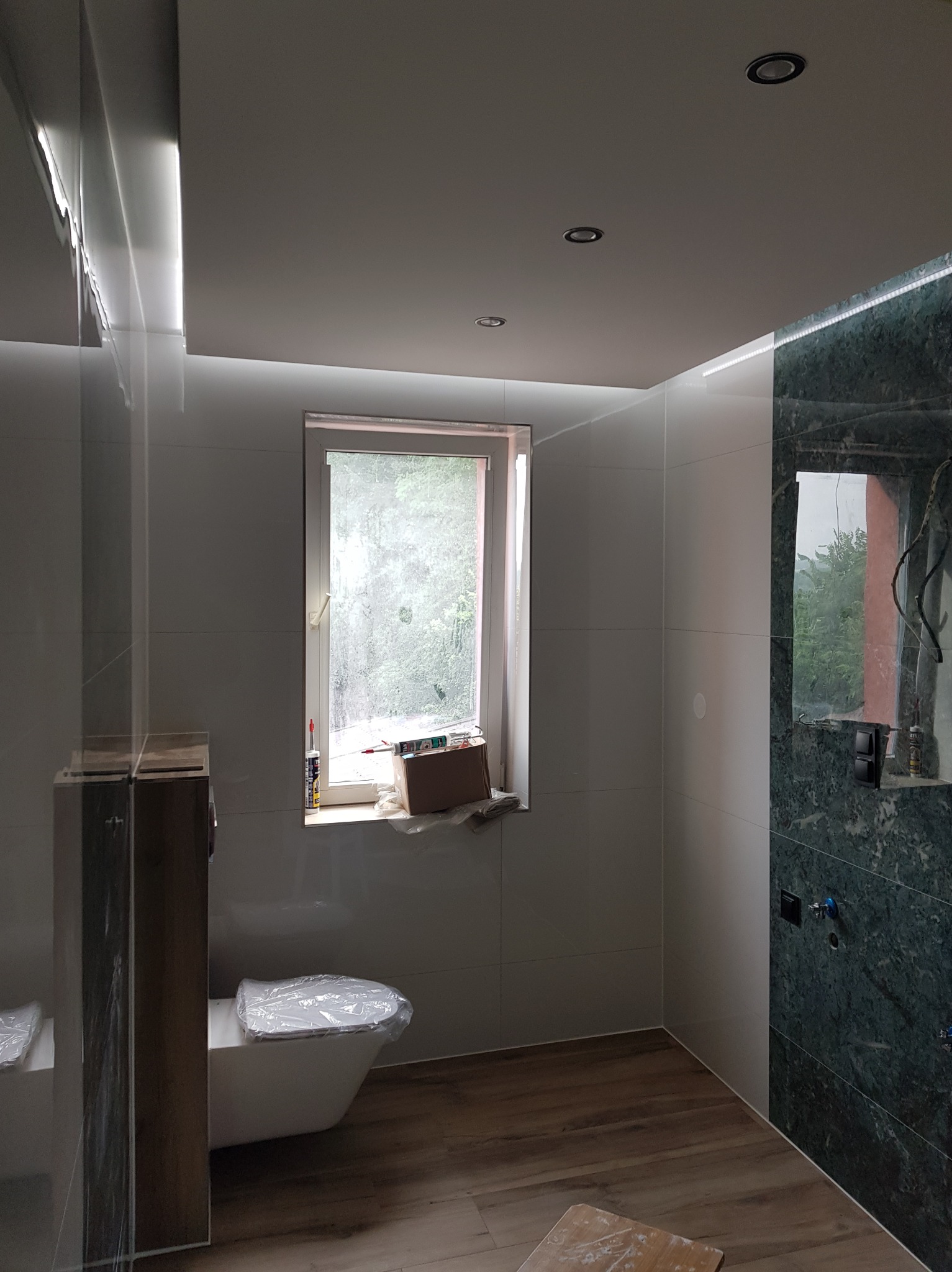 Wykończona łazienka z białymi i zielonymi płytkami, drewnopodobną podłogą i oświetleniem LED. Toaleta zabezpieczona folią, okno z widokiem na zieleń.