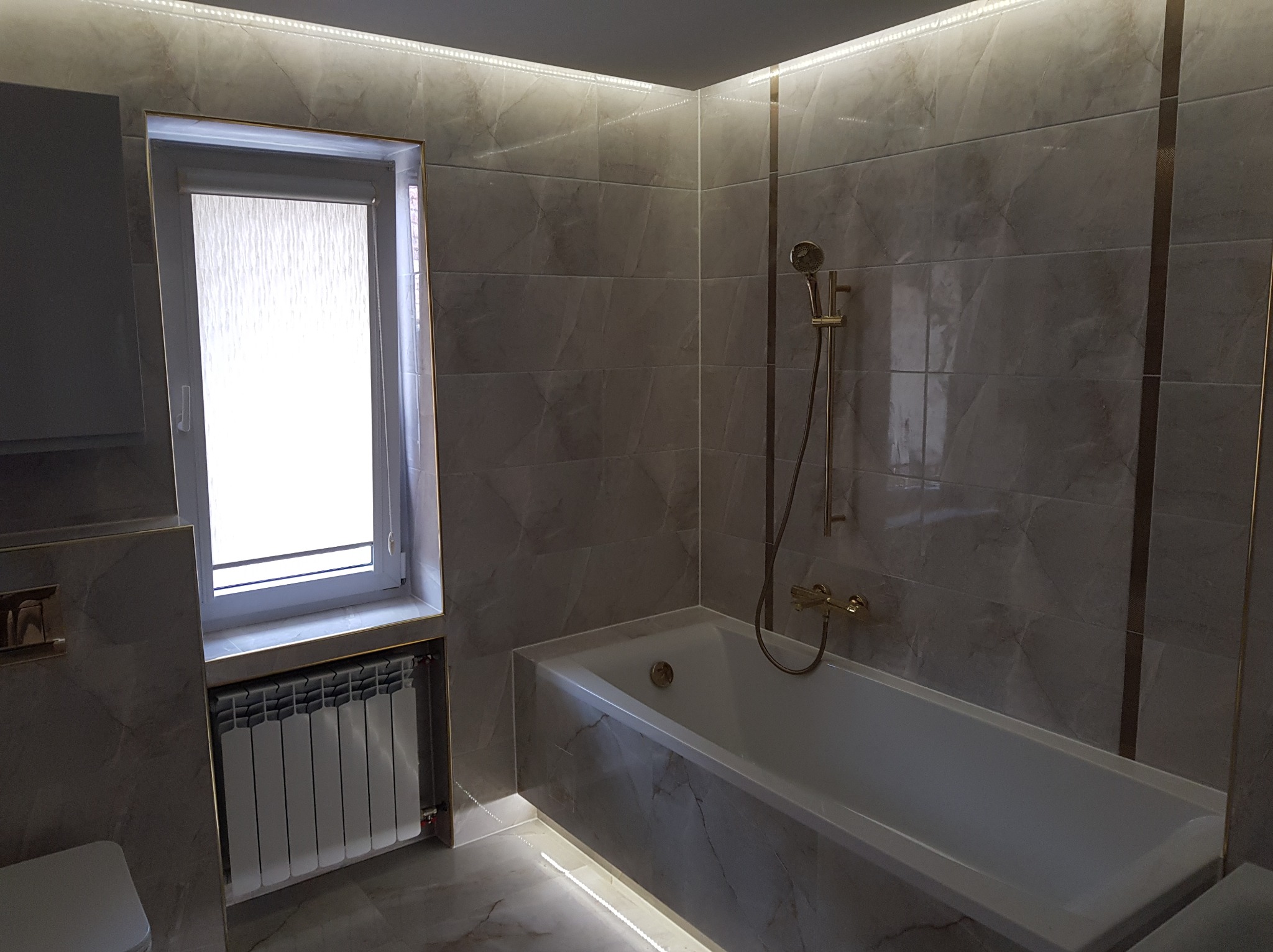 Wykończona łazienka w szarościach z wanną, grzejnikiem pod oknem i oświetleniem LED. Płytki imitujące marmur z akcentami w kolorze złota.