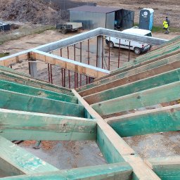 PP. Więźby. - Zielona impregnacja drewnianej więźby dachowej w trakcie budowy domu. Widoczne betonowe fundamenty i elementy szalunkowe. Na zewnątrz pojazd i przenośna toaleta.