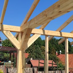 PP. Więźby. - Drewniana konstrukcja altany w trakcie budowy, widoczne elementy nośne i złączenia ciesielskie na tle błękitnego nieba i zieleni drzew. W tle zabudowa jednorodzinna.