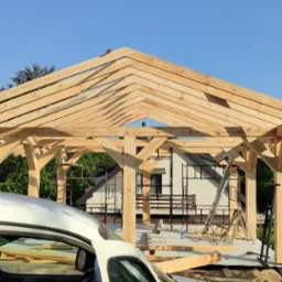 Konstrukcja drewniana altany w trakcie budowy, widoczne elementy więźby dachowej i słupy nośne. W tle budynki mieszkalne i zieleń.