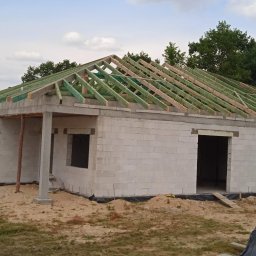 PP. Więźby. - Budowa parterowego domu z bloczków: ściany wzniesione, więźba dachowa w trakcie montażu. Widoczny teren budowy i elementy konstrukcyjne.