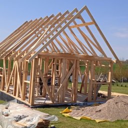 PP. Więźby. - Konstrukcja szkieletowa domu w trakcie budowy, widoczna więźba dachowa na tle zielonego krajobrazu i błękitnego nieba. Etap montażu belek.