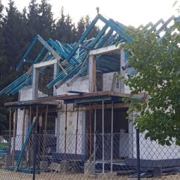 PP. Więźby. - Budowa domu z bloczków: wzniesione ściany parteru, przygotowana więźba dachowa w kolorze niebieskim. W tle las, na pierwszym planie ogrodzenie z siatki.