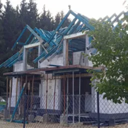 Budowa domu z bloczk&oacute;w: wzniesione ściany parteru, przygotowana więźba dachowa w kolorze niebieskim. W tle las, na pierwszym planie ogrodzenie z siatki.