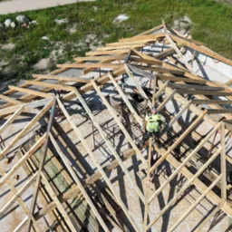 Montaż drewnianej więźby dachowej z perspektywy drona. Dwóch pracowników w trakcie pracy. Uwagę przykuwa precyzja wykonania i jasne drewno konstrukcyjne.