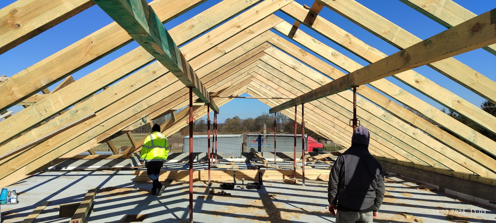 Wnętrze konstrukcji dachu z drewnianą więźbą, widok z dołu na drewniane elementy, podpory i dwóch pracowników w trakcie montażu. Jasne niebo w tle.