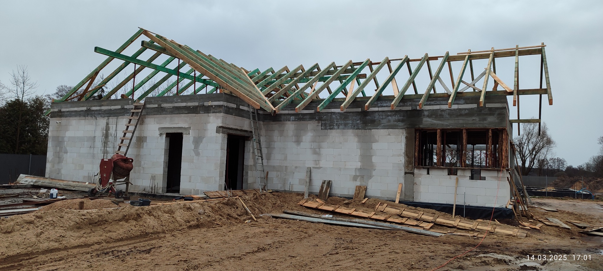 Budowa domu z bloczków: wzniesione ściany i częściowo zamontowana, zielona więźba dachowa. Na pierwszym planie piasek i resztki materiałów budowlanych.