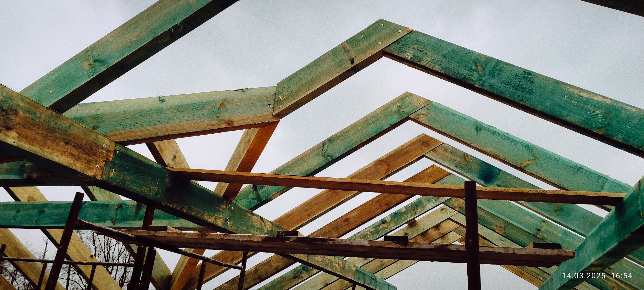 Wznoszenie impregnowanej konstrukcji dachu z drewna, widok z dołu na niebo, Giżycko. Elementy połączone śrubami, widoczne rusztowania i fragmenty drzew.