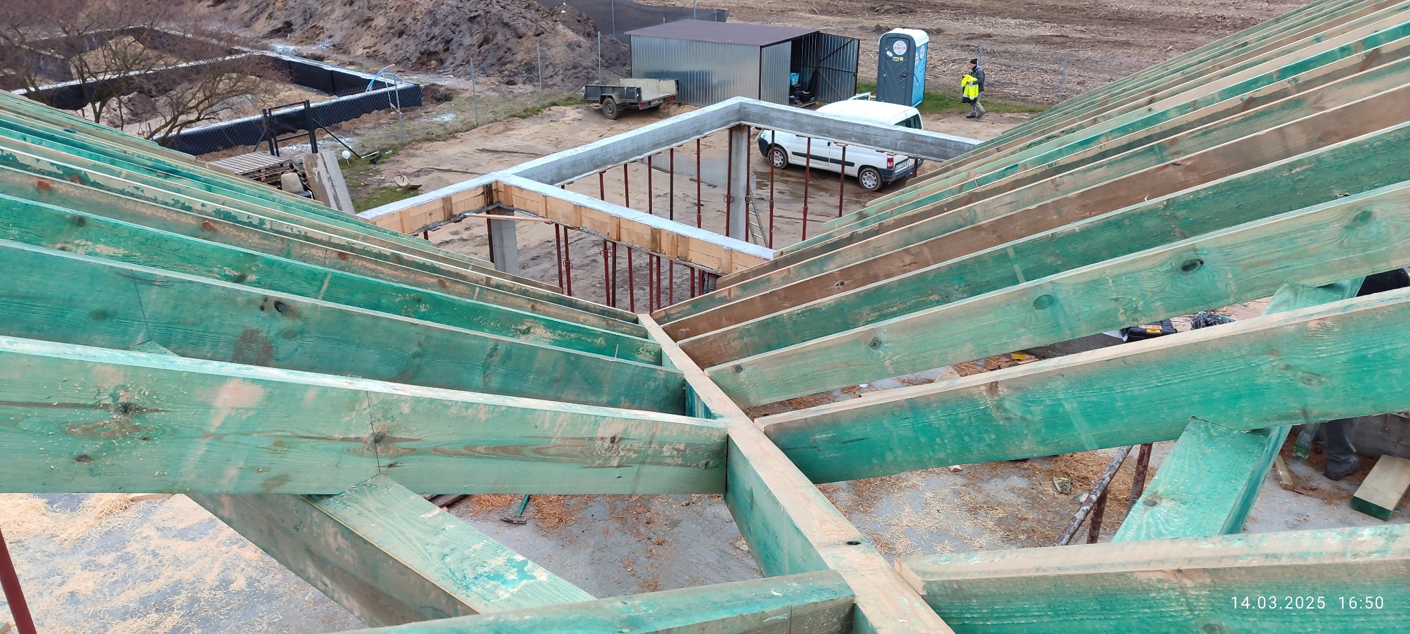 Zielona impregnacja drewnianej więźby dachowej w trakcie budowy domu. Widoczne betonowe fundamenty i elementy szalunkowe. Na zewnątrz pojazd i przenośna toaleta.