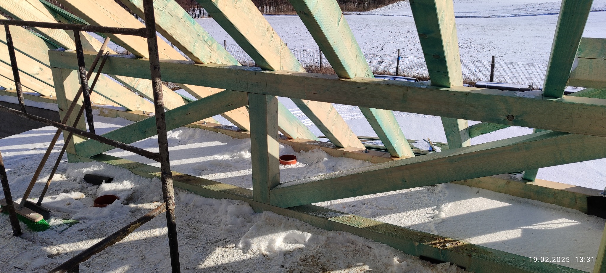 Wnętrze konstrukcji dachu z drewnianymi belkami, widok z góry na ośnieżoną powierzchnię, fragment rusztowania i narzędzia. Prace dekarskie w zimowym krajobrazie.