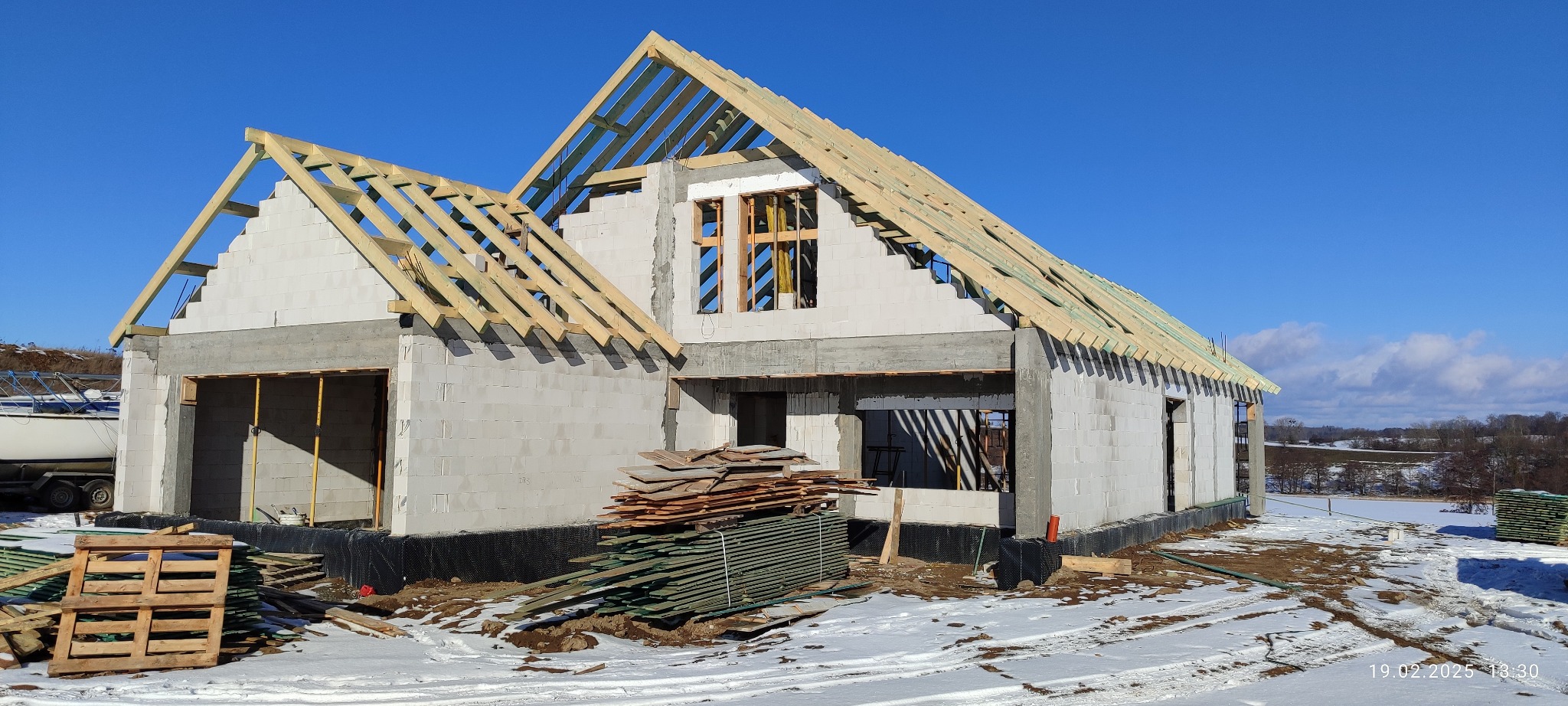 Budowa domu z białych bloczków, widoczna konstrukcja więźby dachowej. Na pierwszym planie deski i paleta, w tle zasnieżony krajobraz i błękitne niebo.