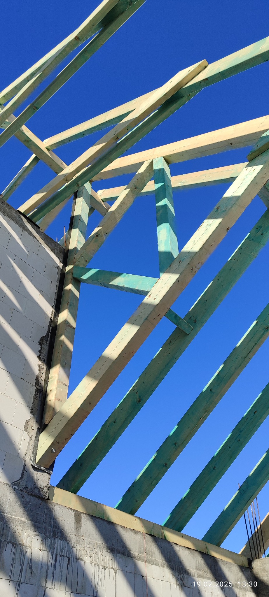 Konstrukcja więźby dachowej z impregnowanego drewna, widok z dołu na tle błękitnego nieba, fragment ściany z betonu komórkowego.