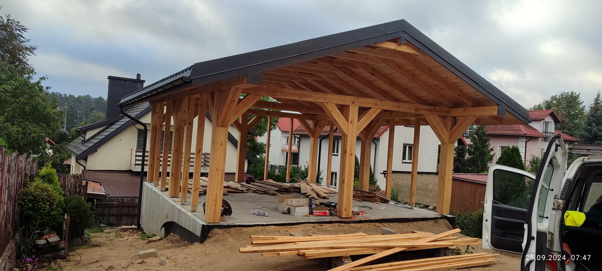 Surowa konstrukcja drewnianej altany z czarnym dachem na betonowej podstawie, widoczna na tle zabudowy mieszkalnej i częściowo otwartego samochodu dostawczego.